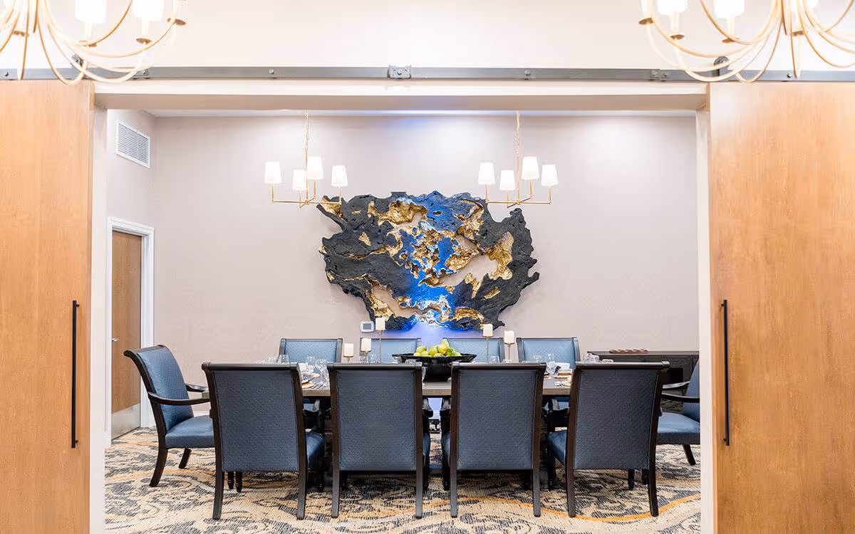 Dining room with a long table surrounded by upholstered chairs, chandeliers overhead, and a large abstract wall sculpture.