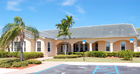 Exterior view of a single-story building with a tiled roof, beige walls, and several windows. The entrance is covered by an archway supported by columns. There are palm trees and shrubs in front of the building, with a clear blue sky above. Handicap parking spaces are visible in the foreground.
