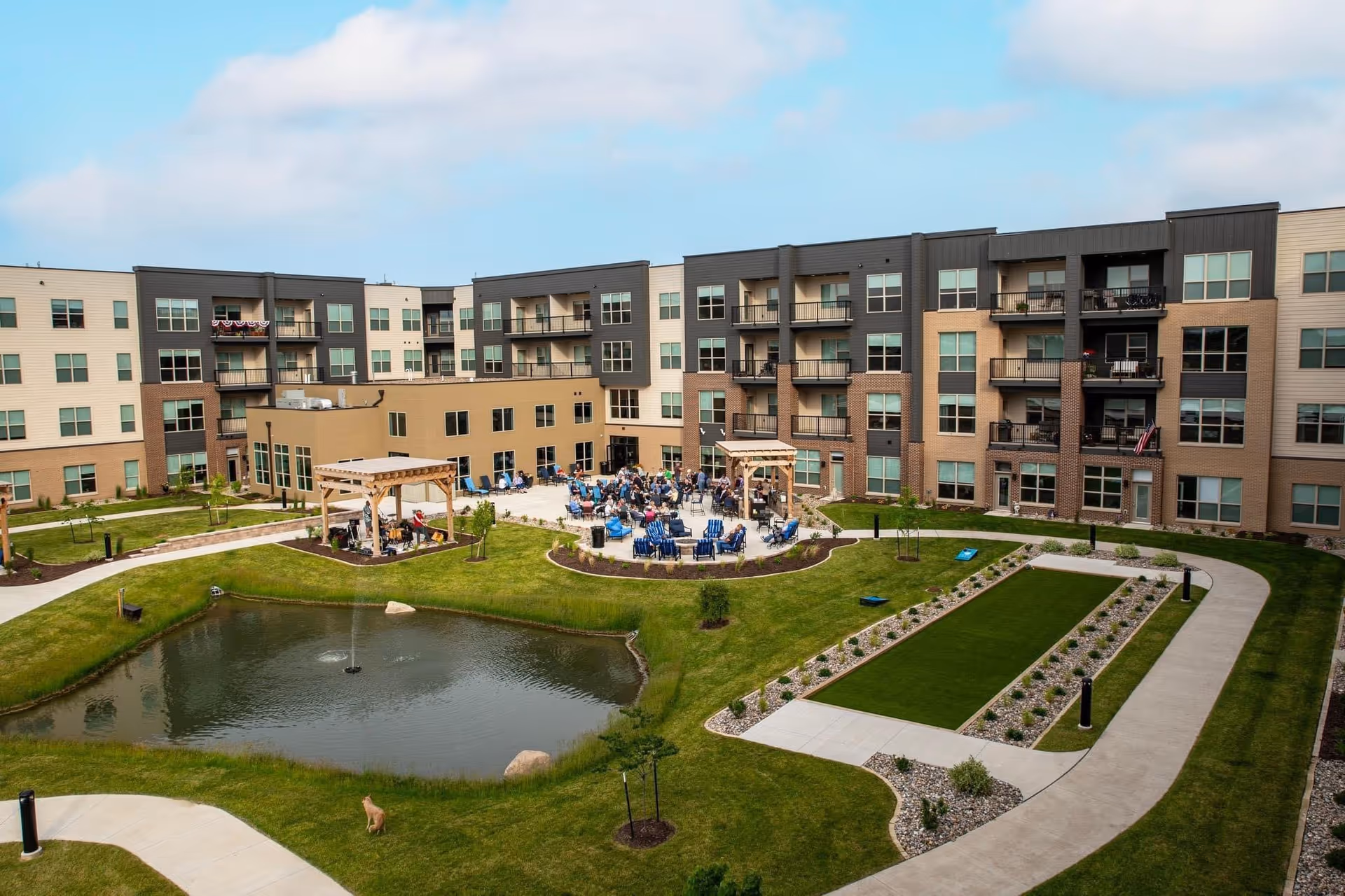 Outdoor courtyard area of a senior living facility with a small pond featuring a fountain, green lawns, walking paths, and a gathering of people seated around tables under pergolas. The building surrounds the courtyard with multiple floors and balconies.
