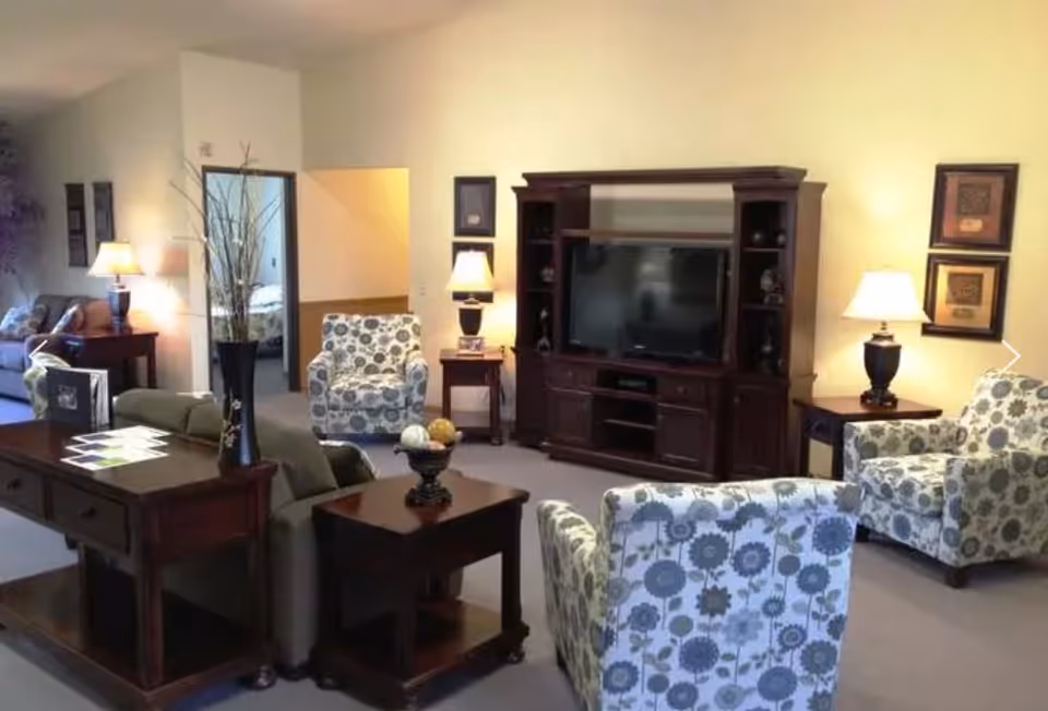 A cozy living room area with patterned armchairs, a sofa, wooden side tables with lamps, a large wooden entertainment center with a flat-screen TV, and a vase with decorative branches on a table. There is an open doorway leading to a bedroom in the background.