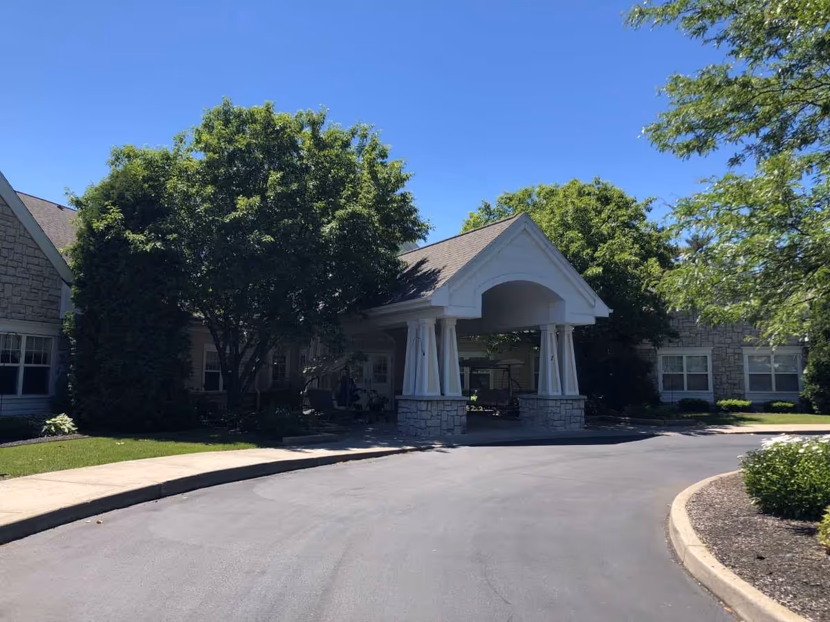 Entrance to a senior living facility with a covered drop-off area supported by white columns and stone bases. The building exterior is made of stone and siding, surrounded by green trees and landscaping under a clear blue sky.