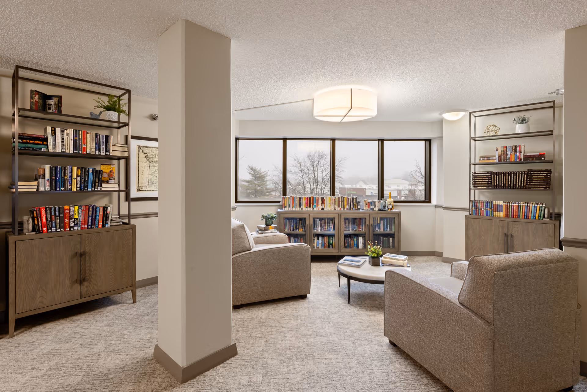 A cozy common room with armchairs, a low coffee table and bookshelves in front of large windows.