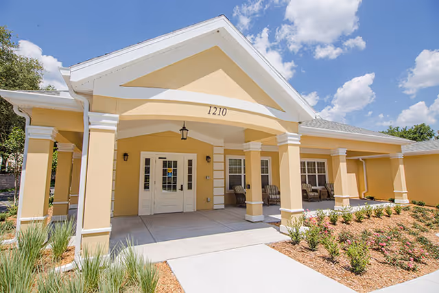 Front entrance of a yellow senior living facility with a covered portico, double doors, and landscaped walkway.