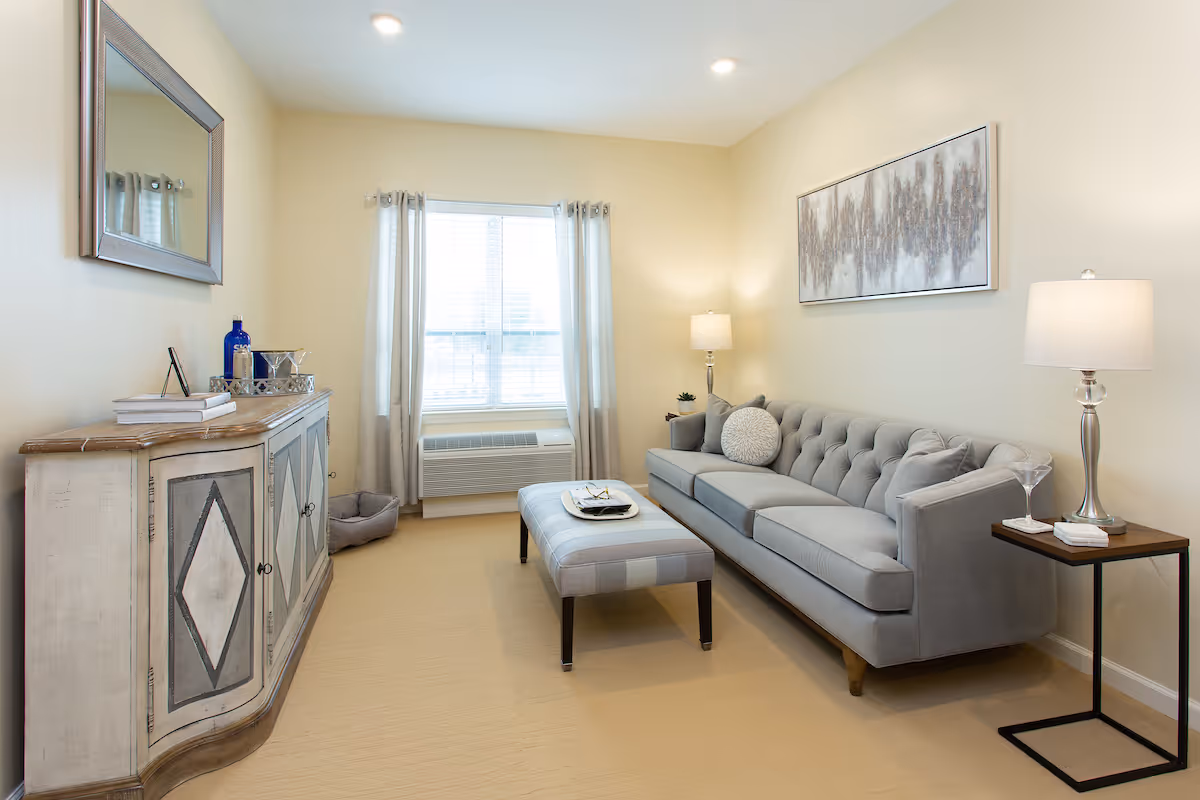 A cozy living room with a light gray tufted sofa adorned with round and square pillows, a matching ottoman in front of it, a wooden side table with a lamp and a glass, and a decorative cabinet with a mirror above it. The room has beige walls, a window with light curtains, and soft lighting from ceiling lights and lamps.