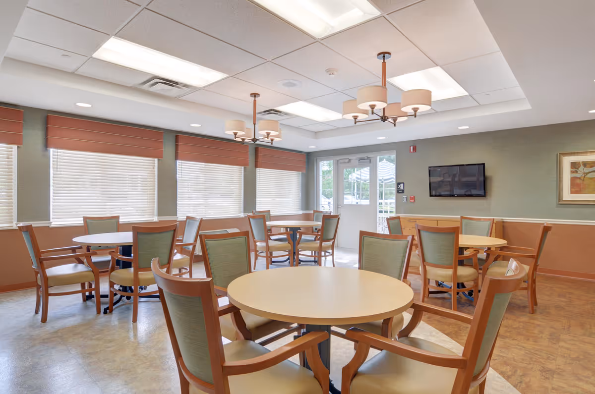 Bright dining room with round tables and wooden chairs, ceiling lights, windows with blinds, and a wall-mounted TV.