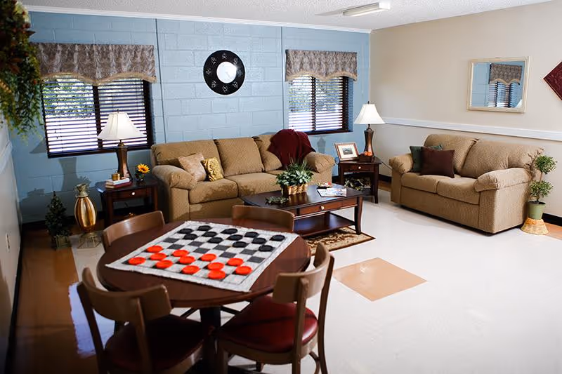Well-lit assisted living common room with two sofas, a coffee table, and a small round table set up with a checkers board.