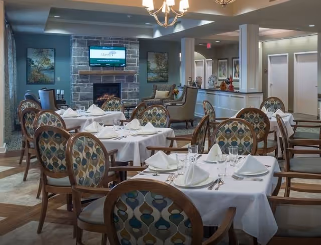 A dining room with several tables covered in white tablecloths, each set with folded napkins, glasses, and silverware. The chairs have patterned upholstery. In the background, there is a stone fireplace with a TV mounted above it, flanked by two framed paintings. The room has soft lighting and a cozy atmosphere.