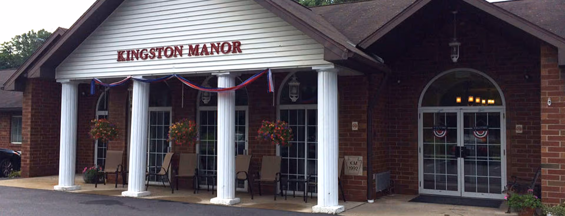 Exterior front entrance of Kingston Manor, a brick building with white columns and a covered porch. There are chairs and hanging flower baskets on the porch, and red, white, and blue bunting decorations hanging above the columns.