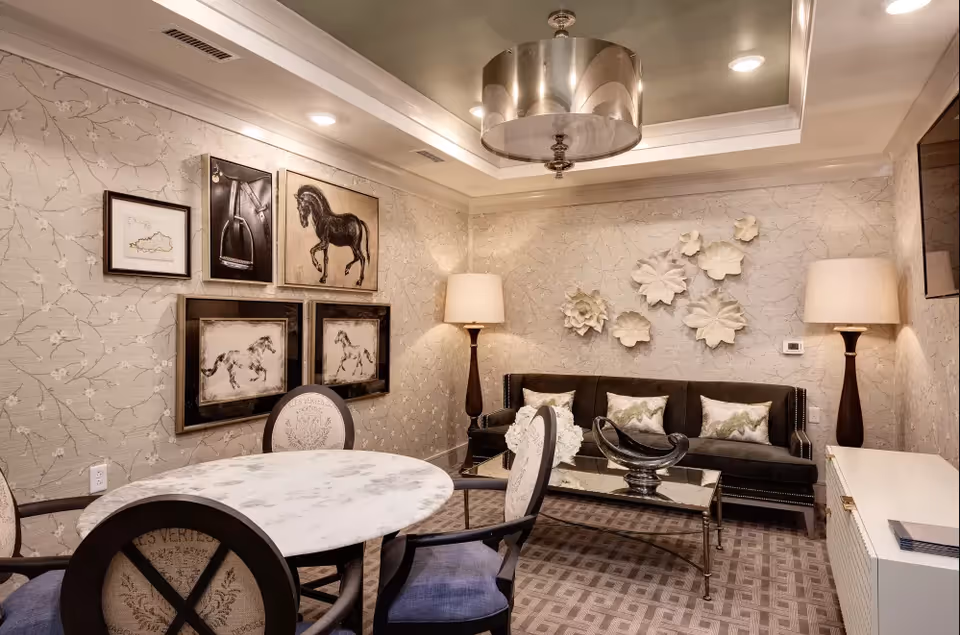 A cozy living room area with a round marble table and four upholstered chairs in the foreground. The walls are decorated with framed horse artwork and white floral wall sculptures. There is a dark sofa with decorative pillows, two tall floor lamps with beige shades, a glass coffee table with a decorative bowl, and a patterned carpet. The ceiling has recessed lighting and a modern chandelier.