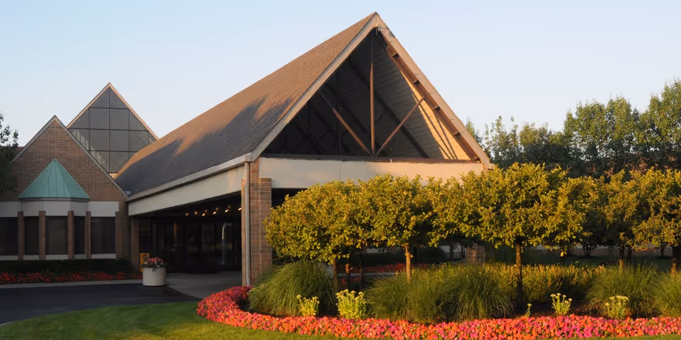 Front entrance of a building with a steep triangular roof, covered driveway, and manicured flower beds and shrubs.