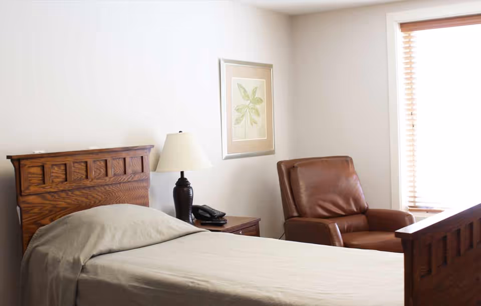 Simple bedroom with a wooden single bed, nightstand with a lamp and phone, and a leather armchair by a window.