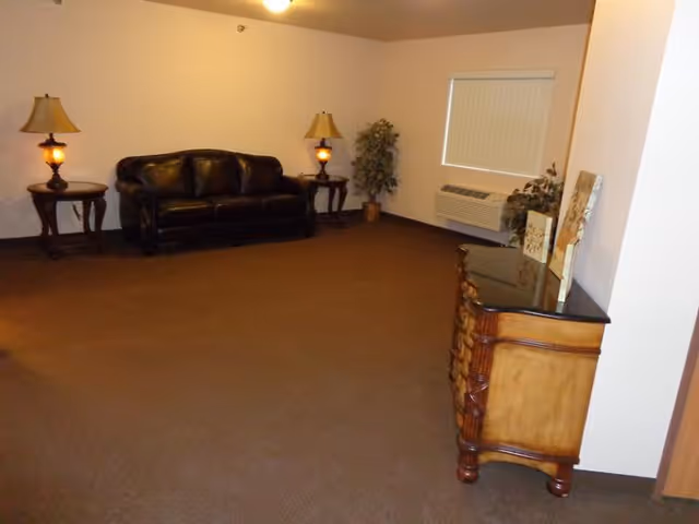 A simple living room with a dark brown leather sofa centered against a beige wall, flanked by two wooden side tables each with a lamp. There is a potted plant in the corner near a window with closed blinds and an air conditioning unit below it. A wooden dresser with a dark top is positioned on the right side of the room with some decorative items on it. The floor is carpeted in brown.