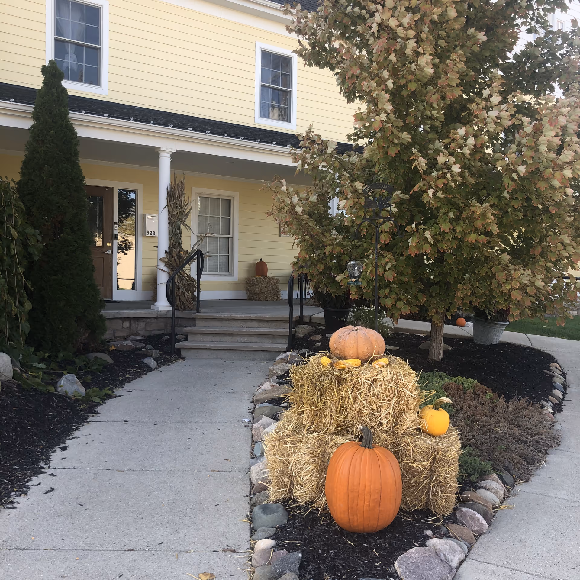 Front entrance of a yellow building with white columns and a brown door. The walkway is lined with rocks and mulch, and there are autumn decorations including hay bales, pumpkins, and corn. A tree with green and brown leaves is on the right side of the entrance.
