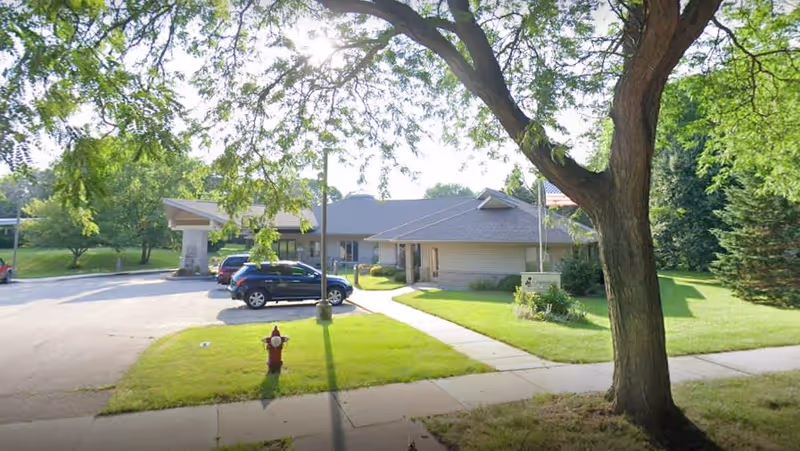 Exterior view of Chamomile Assisted Living Ltd facility showing a single-story building with a gray roof, surrounded by green grass and trees. There are several parked cars in the driveway and a sidewalk leading to the entrance. A large tree is in the foreground on the right side.