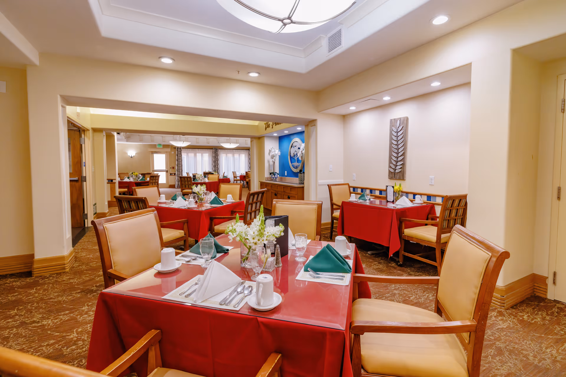 Dining room with multiple tables covered in red tablecloths, place settings and floral centerpieces under warm ceiling lighting.