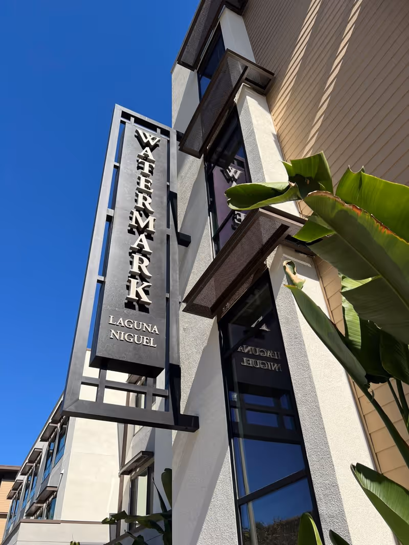 Exterior view of a modern building with a vertical sign that reads 'Watermark Laguna Niguel' against a clear blue sky, with green tropical plants in the foreground.
