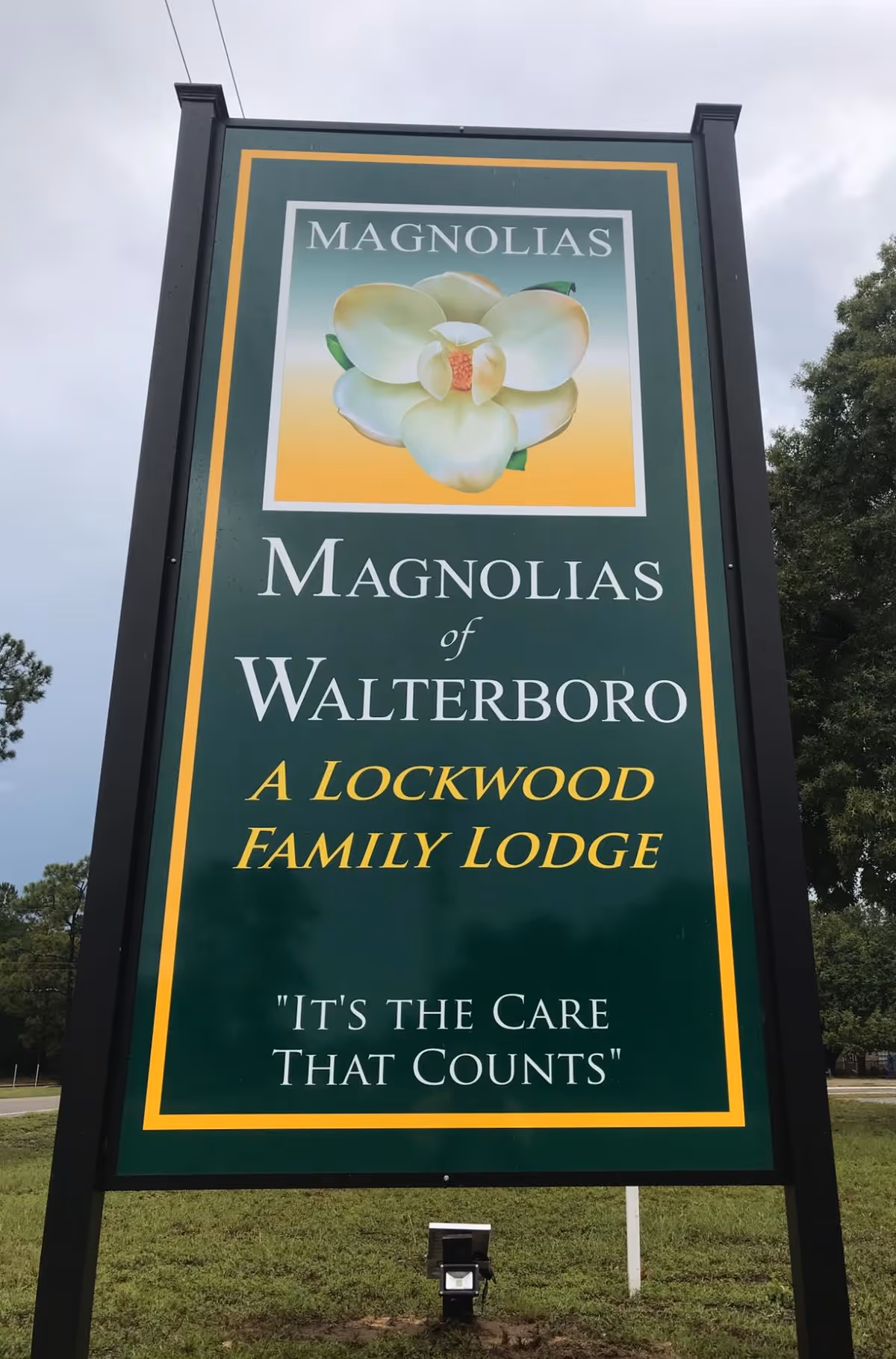 A large green entrance sign for "Magnolias of Walterboro" with a magnolia flower logo and the slogan "It's the Care That Counts."