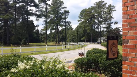 Brick building entrance sign beside a curved road, white fence, and tall pine trees outdoors.