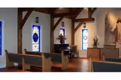 Interior view of a small chapel with wooden pews, stained glass windows, wooden beams on the ceiling, a piano, and a religious painting on the wall behind an altar.