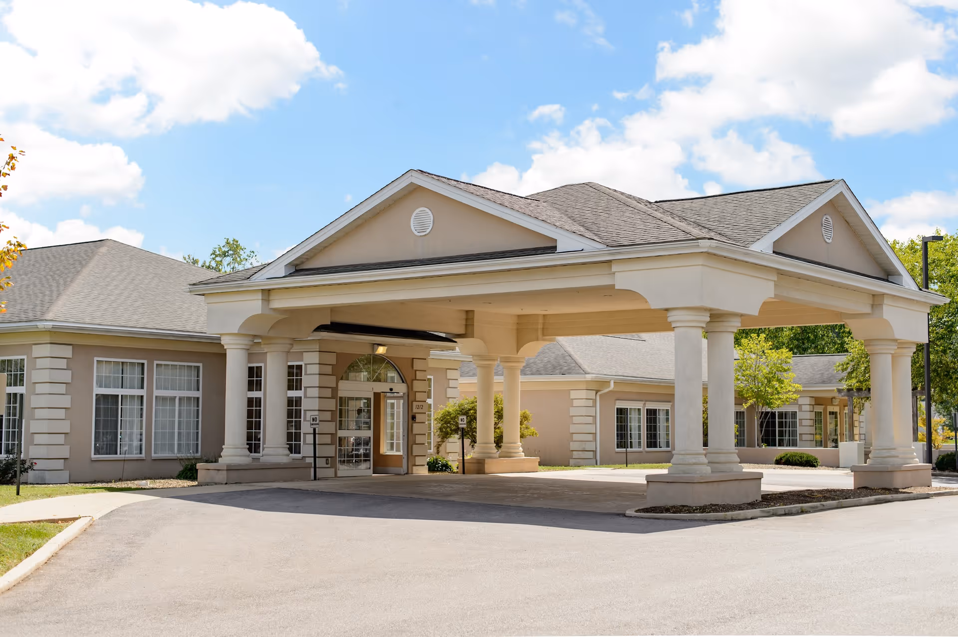 Front exterior view of a single-story senior living facility building with a covered entrance supported by large columns, surrounded by a paved driveway and some greenery under a partly cloudy sky.