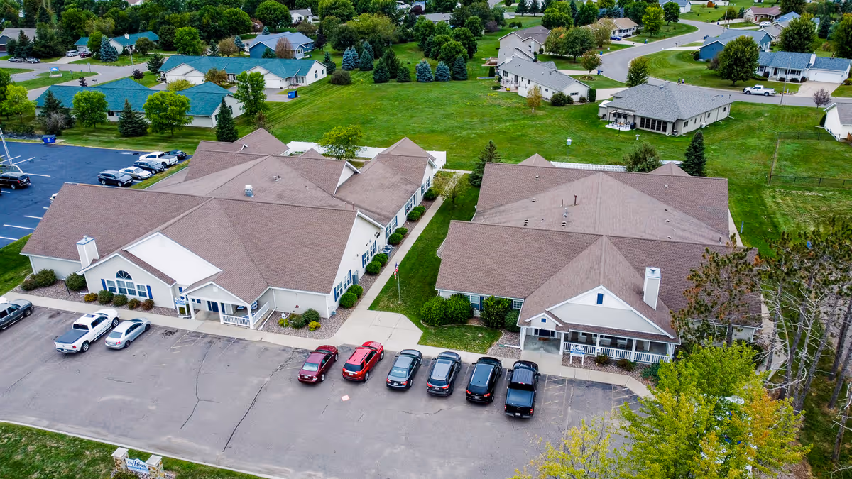 Aerial view of a senior living facility with two large connected buildings surrounded by green lawns and trees. Several cars are parked in front of the buildings, and residential houses are visible in the background.