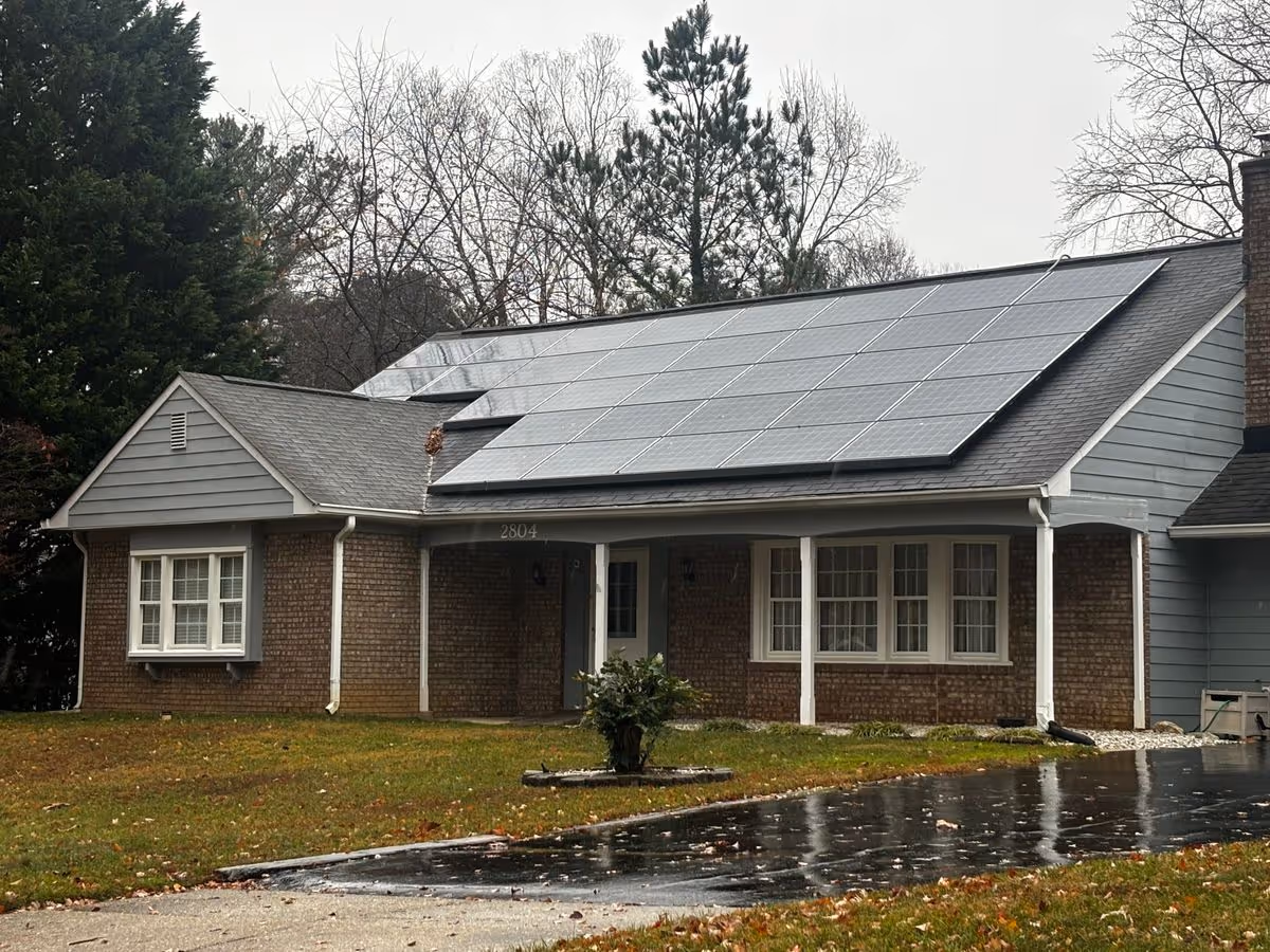 Single-story brick house front with a covered porch and large solar panels on the roof and a wet driveway in the foreground.