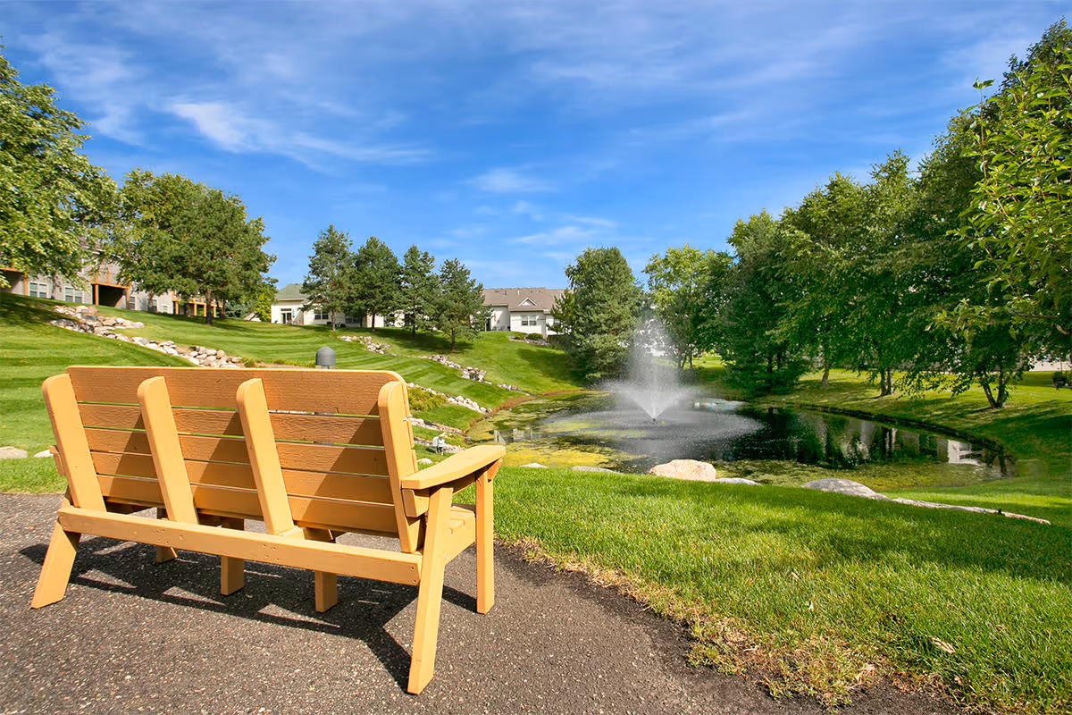 A wooden bench on a paved path overlooks a small pond with a fountain in the middle, surrounded by green grass, trees, and residential buildings under a blue sky with some clouds.