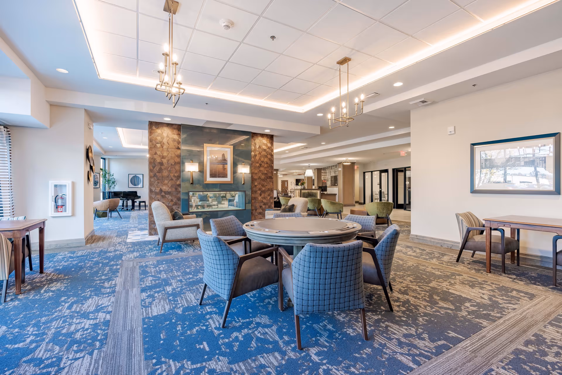 A spacious and well-lit common area in a senior living facility featuring a round table with six upholstered chairs in the foreground. The room has a modern design with blue and beige patterned carpet, two chandeliers hanging from a recessed ceiling with LED lighting, and a decorative fireplace with artwork above it. In the background, there are additional seating areas including green chairs and a piano near the windows.