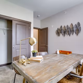 A rustic wooden dining table with chairs, a lamp, books, and decorative wall art in a bright apartment room showing an open closet and doorway.