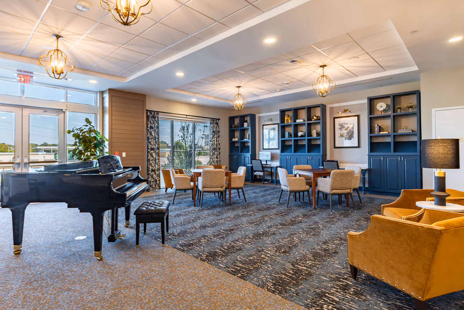 A spacious and well-lit common area in a senior living facility featuring a black grand piano with a matching bench on the left, several round tables with beige upholstered chairs, built-in blue shelving units with decorative items and framed artwork on the back wall, and two mustard yellow armchairs with a small round table and lamp on the right. Large windows and glass doors allow natural light to fill the room.
