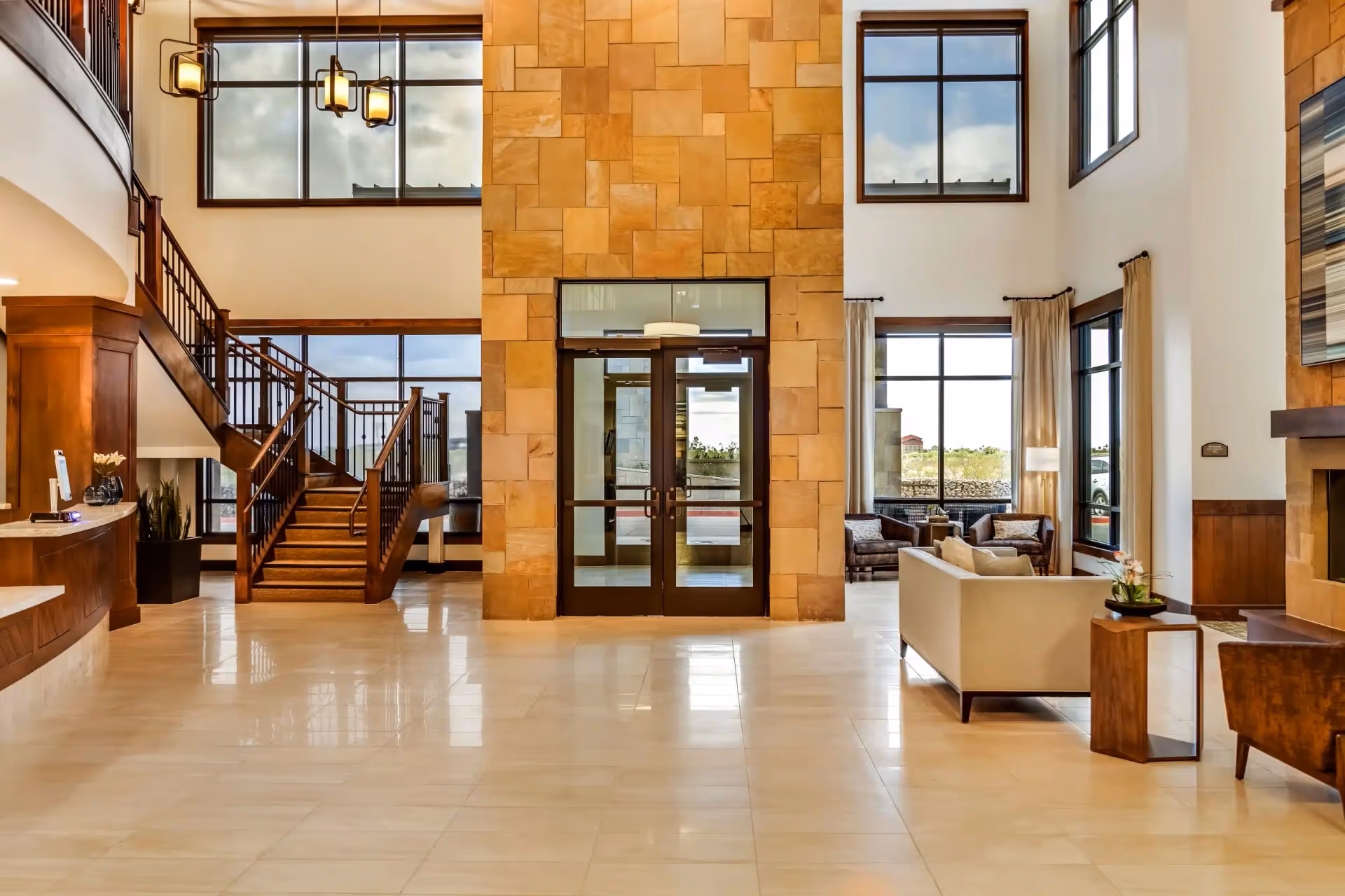 Spacious and well-lit lobby area with large windows, a central stone wall with double glass doors, a wooden staircase on the left, and a seating area with sofas and chairs on the right.