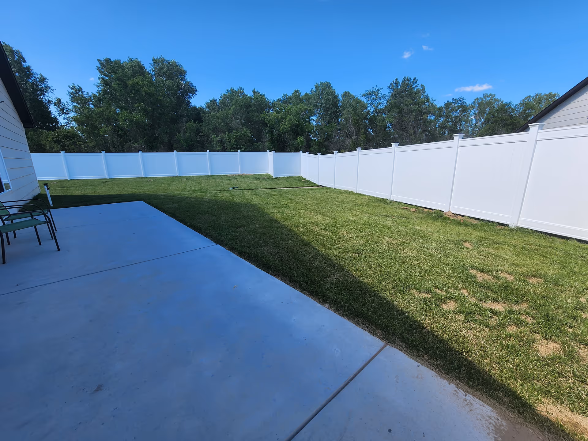 Outdoor view of a fenced backyard with a concrete patio area and green grass lawn, surrounded by a white privacy fence and trees in the background under a clear blue sky.