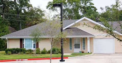 Single-story beige senior living facility with shrubs, a driveway, and a centered entrance.