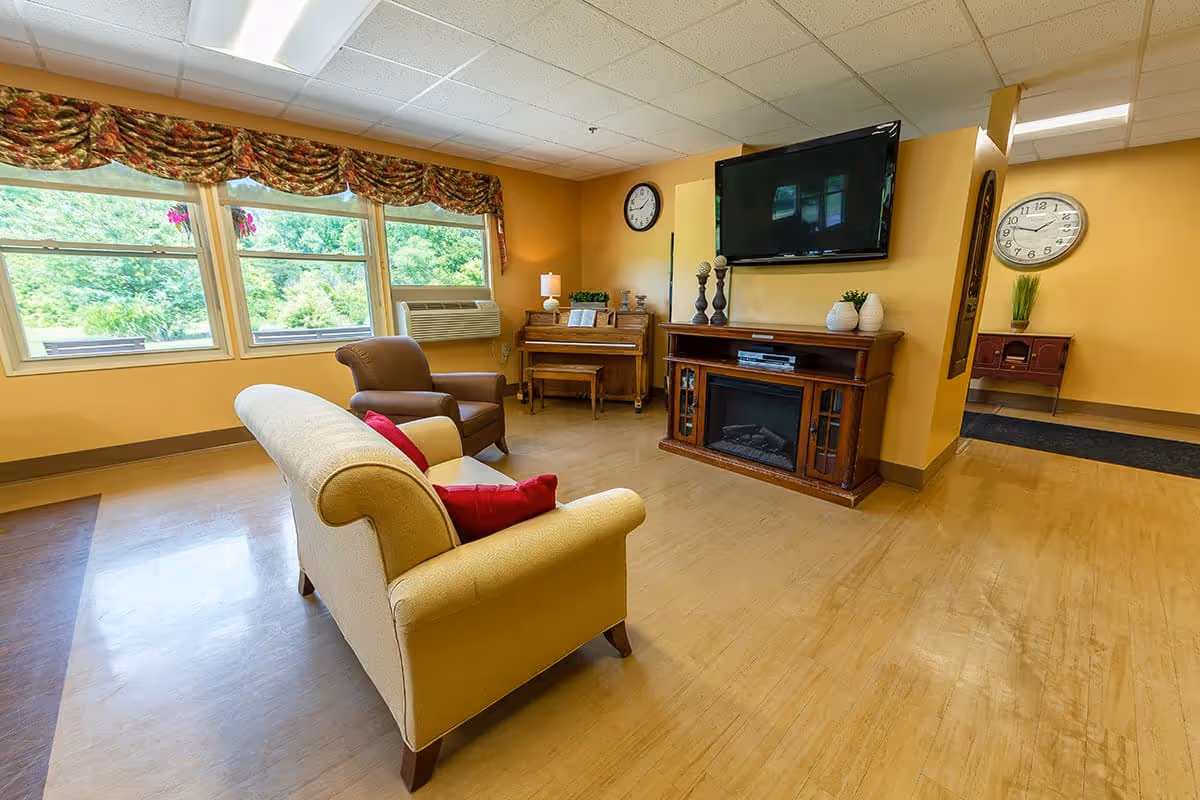 A cozy living room area with a beige couch adorned with red pillows, a brown armchair, a wooden piano with a bench, and a wooden entertainment center with a flat-screen TV mounted above it. The room has large windows with floral valances letting in natural light, a wall clock, and light yellow walls with a wooden floor.