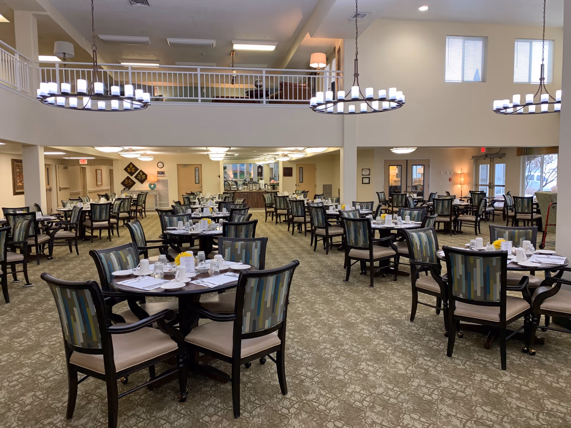 Spacious senior living dining room with multiple round tables set for meals, patterned chairs, and hanging chandeliers.