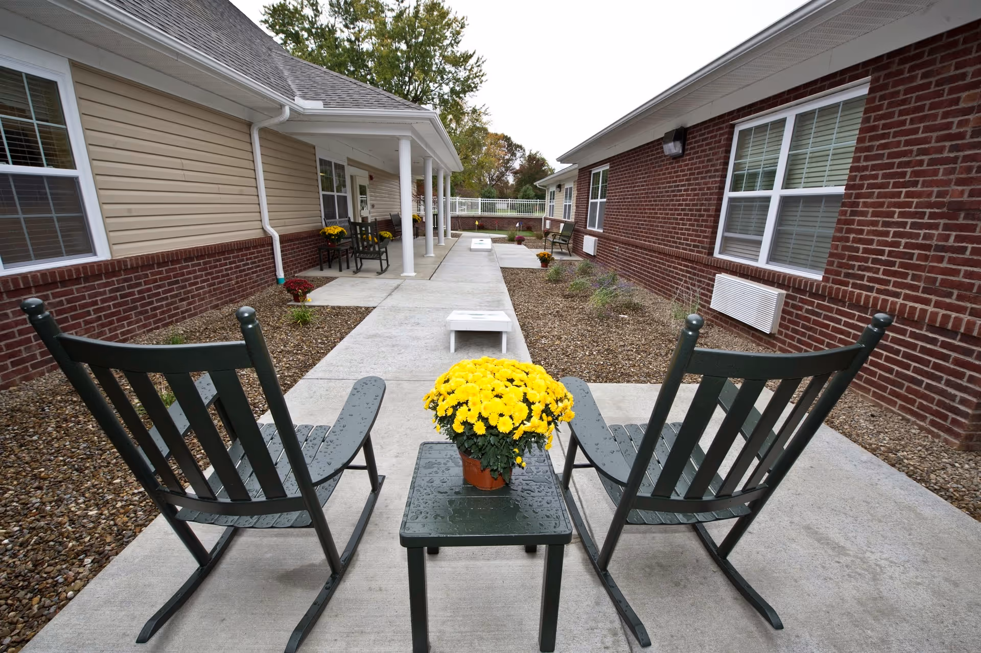 Outdoor patio area between two single-story buildings with brick and beige siding. The patio has a concrete walkway with two green rocking chairs and a small table holding a pot of yellow flowers in the foreground. There are additional chairs and tables along the walkway, and some plants and mulch beds on either side.