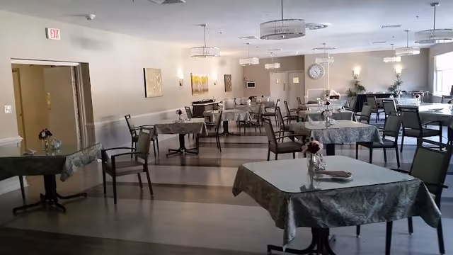 A spacious dining room with multiple tables covered in patterned tablecloths, each table set with a small floral centerpiece. The room has wooden flooring, several chairs around each table, wall decorations, and ceiling lights. A large clock is visible on the far wall, and natural light enters through windows on the right side.