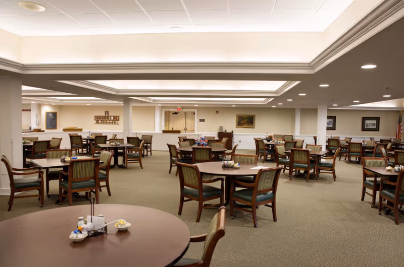 Spacious dining room with multiple round tables and wooden chairs under recessed ceiling lighting.