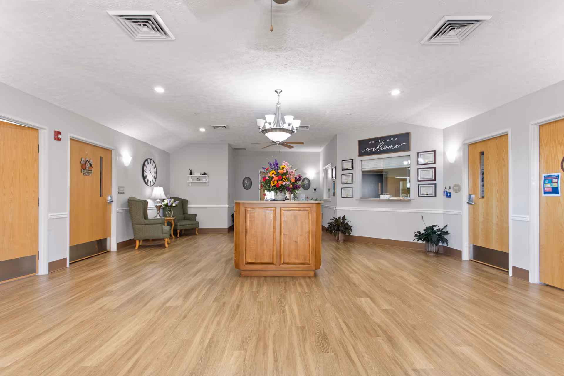 Interior view of a senior living facility lobby area with wooden flooring, a central wooden reception desk adorned with a colorful flower arrangement, two green armchairs with a small table and lamp on the left side, several wooden doors along the walls, a clock on the left wall, and a reception window with a welcome sign and framed certificates on the right wall.