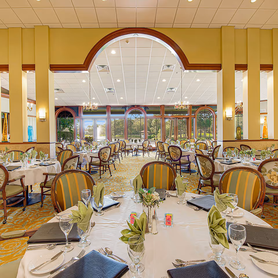 Elegant dining room with round tables covered in white tablecloths, set with glassware, silverware, and green folded napkins. The room features striped and floral upholstered chairs, large arched windows letting in natural light, and chandeliers hanging from the ceiling.