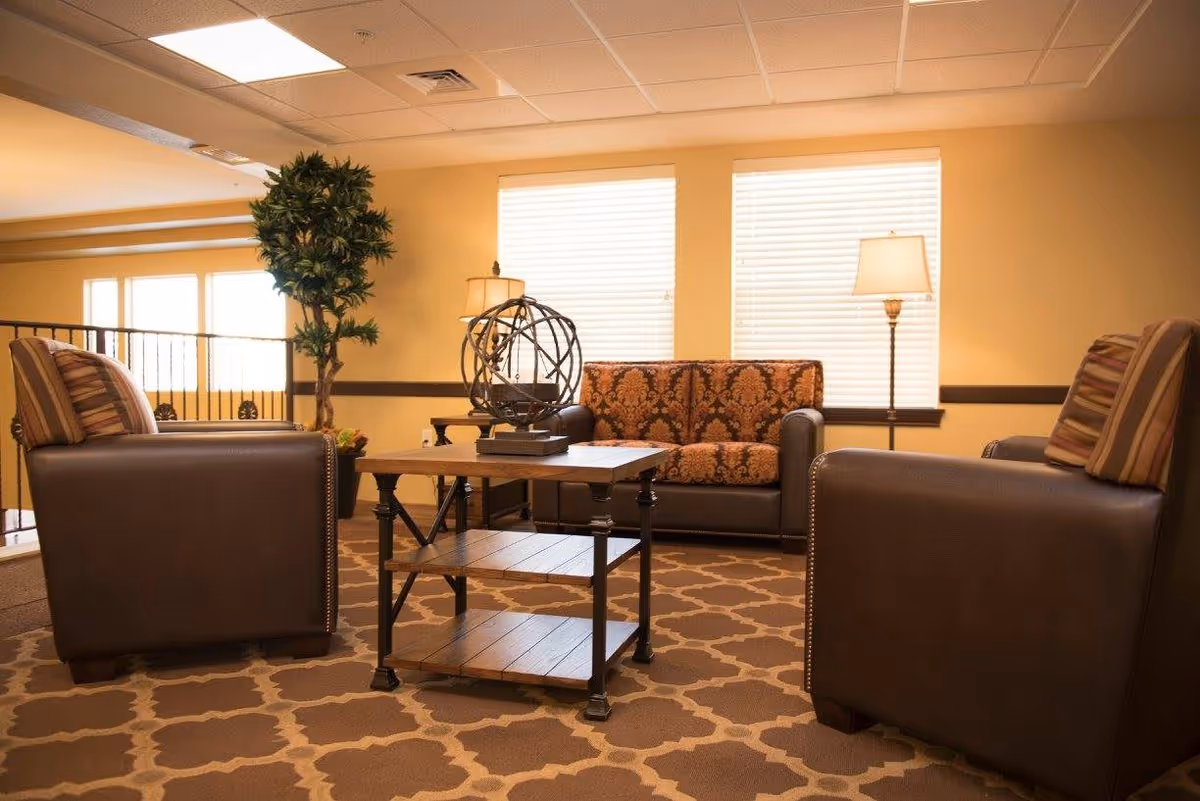 A cozy senior living common area with two brown leather armchairs and a patterned sofa arranged around a wooden coffee table. The room has beige walls, two large windows with blinds, a floor lamp, a table lamp, a decorative plant, and a patterned carpet.