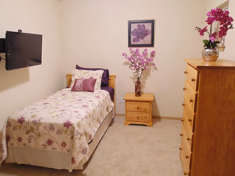 A small bedroom with a single bed covered in a floral bedspread with purple and beige tones. There are three pillows on the bed, including a decorative purple one. A wooden nightstand next to the bed holds a vase with purple flowers. Above the nightstand, a framed picture of a purple flower hangs on the wall. On the right side of the room, there is a tall wooden dresser with a silver vase containing purple orchids on top. A flat-screen TV is mounted on the wall to the left of the bed. The room has beige walls and carpeted flooring.