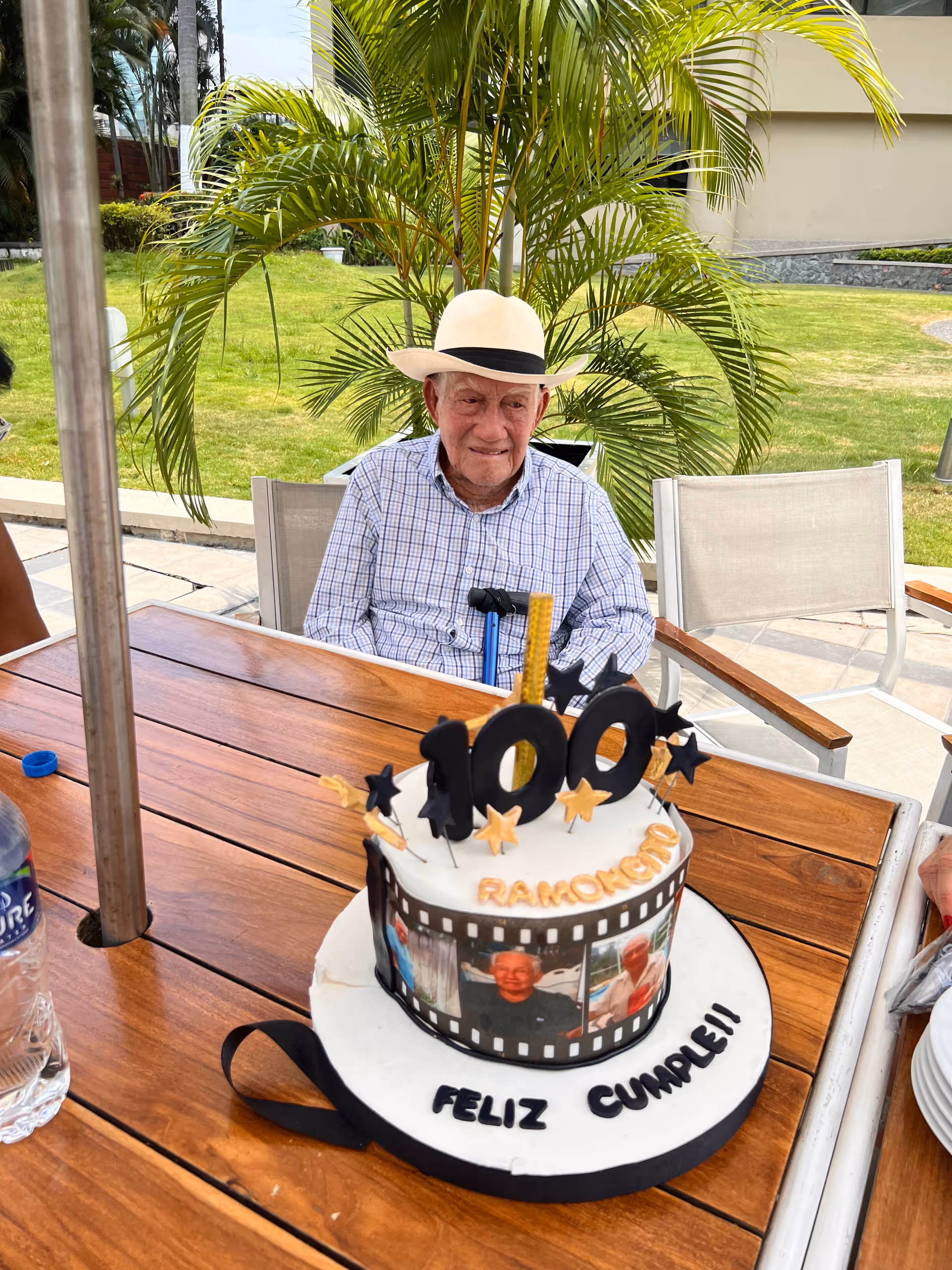 An elderly man wearing a white hat and checkered shirt sits at a wooden outdoor table with a birthday cake in front of him. The cake is decorated with the number 100, stars, and photos, with the words 'RAMONCITO' and 'FELIZ CUMPLE!!' written on it. There is a green lawn and palm plants in the background.