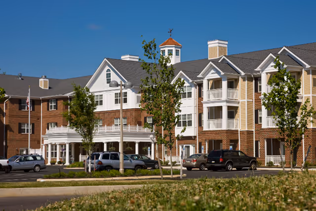 Exterior view of a multi-story senior living facility building with brick and white siding, several windows, balconies, and a cupola on the roof. There are trees and parked cars in front of the building under a clear blue sky.