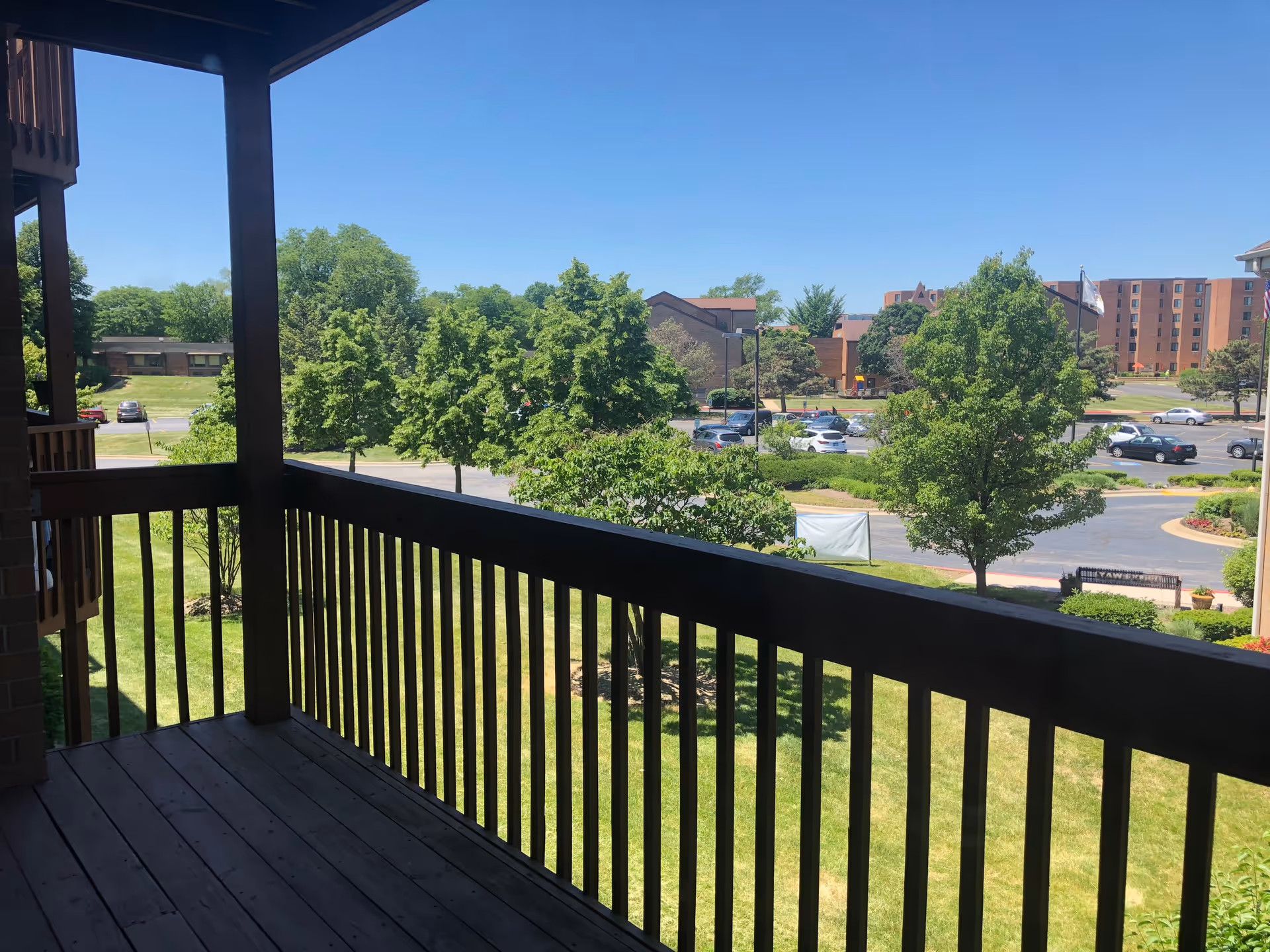 View from a wooden balcony overlooking a grassy area with trees, a parking lot with several cars, and buildings in the background under a clear blue sky.