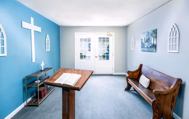 A small chapel room with a wooden podium holding an open book, a wooden bench with a cushion, a blue accent wall with a large white cross and two window-shaped wall decorations, a small shelf with books and a small cross, and double glass doors letting in natural light.