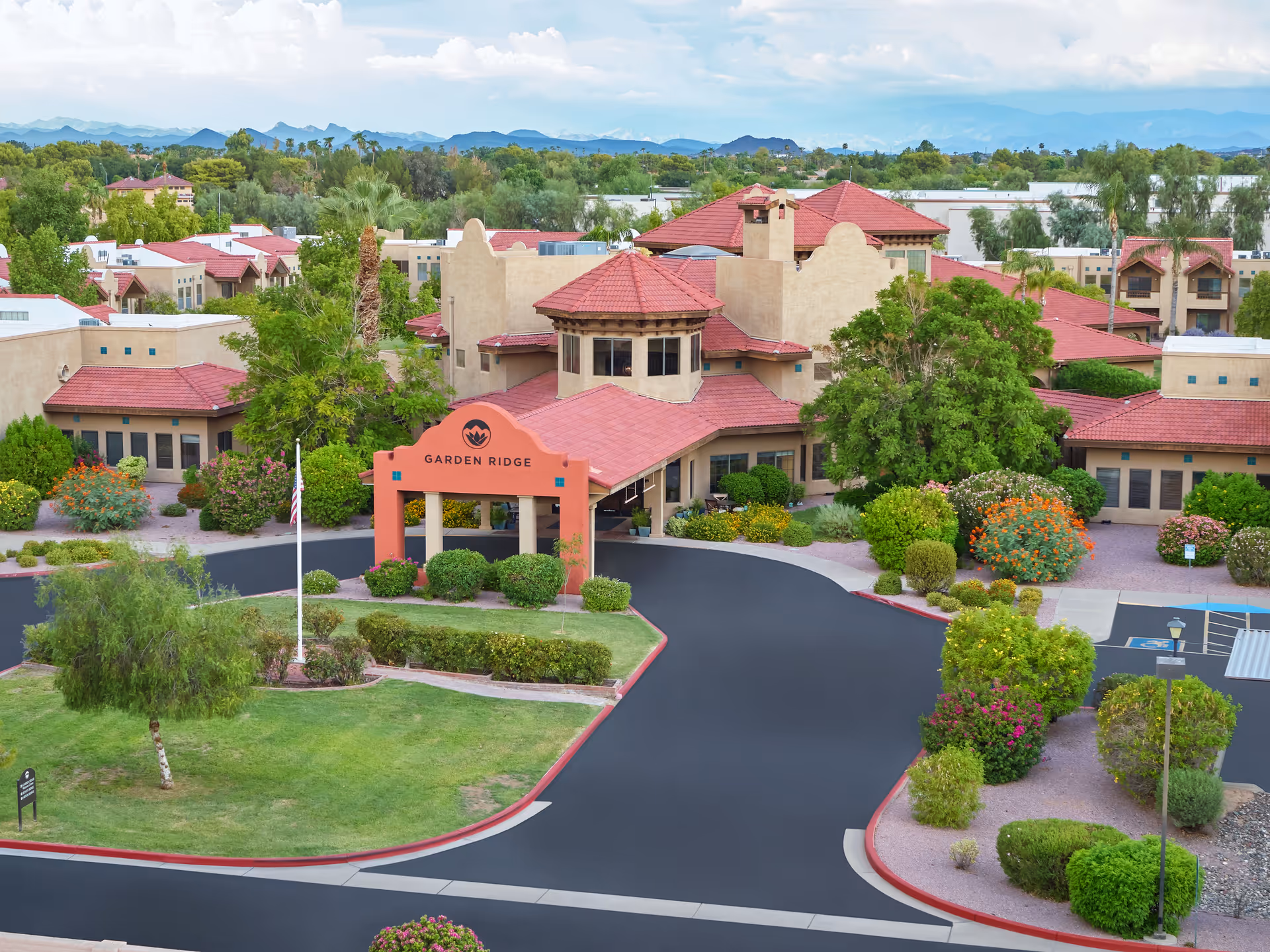 Aerial view of a senior living facility named Garden Ridge with beige stucco buildings and red tile roofs surrounded by green trees, bushes, and landscaped gardens. The driveway curves around a grassy area with a flagpole, and mountains are visible in the background under a partly cloudy sky.