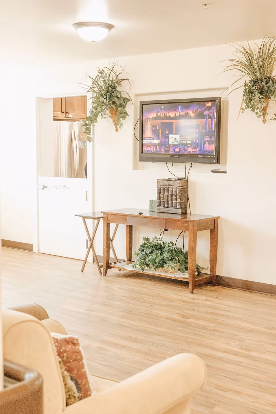 A cozy living room area with a wall-mounted flat-screen TV displaying a colorful cityscape. Below the TV is a wooden console table with decorative books and a green plant underneath. Two hanging plants are mounted on the wall on either side of the TV. A door to the left leads to a kitchen area with a stainless steel refrigerator visible. The floor is light wood, and a beige armchair with a patterned pillow is partially visible in the foreground.