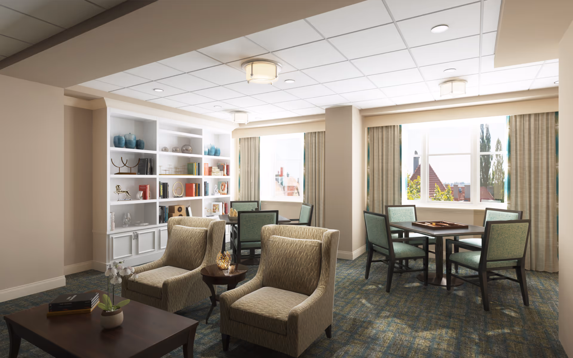 A bright and cozy common area with two beige armchairs and a small round table between them in the foreground. Behind them are two square tables each surrounded by four green upholstered chairs. A large white built-in bookshelf filled with decorative items and books is against the left wall. Large windows with beige curtains allow natural light to fill the room.