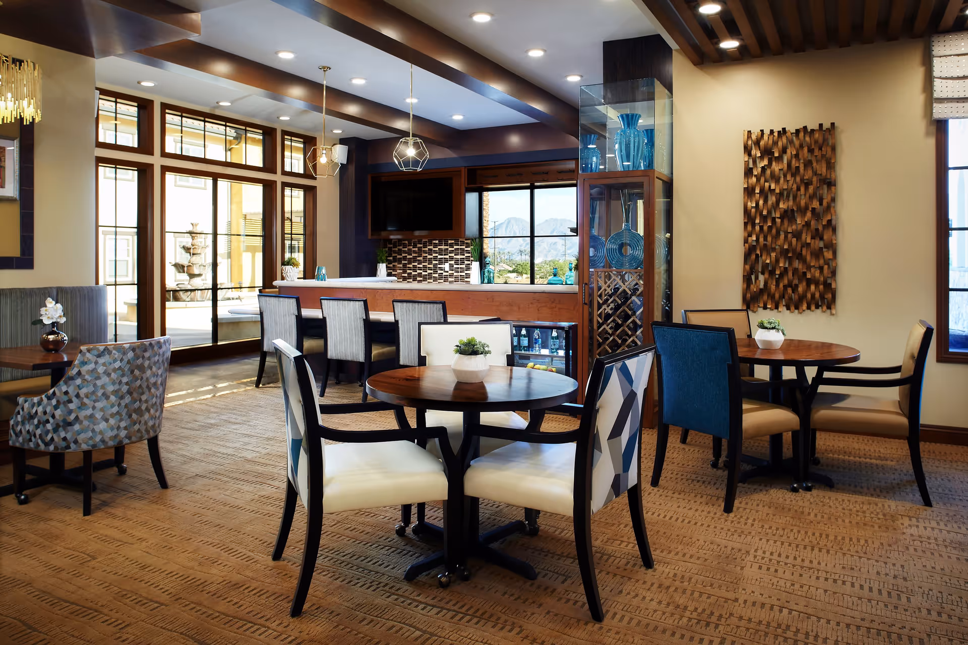 A cozy dining area in Bella Villaggio featuring round wooden tables with cushioned chairs, a bar counter with high chairs, large windows allowing natural light, decorative wall art, and a display cabinet with blue glass vases.